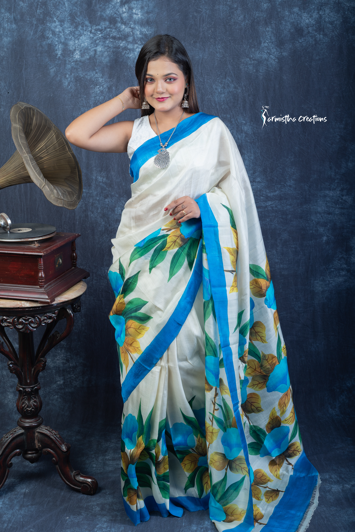 This floral hand painted Bishnupuri silk saree features vibrant blue blooms and mustard-gold leaves on a soft ivory base with a sky-blue border. Made from pure Bengal silk, it’s graceful, light, and perfect for radiant daywear.