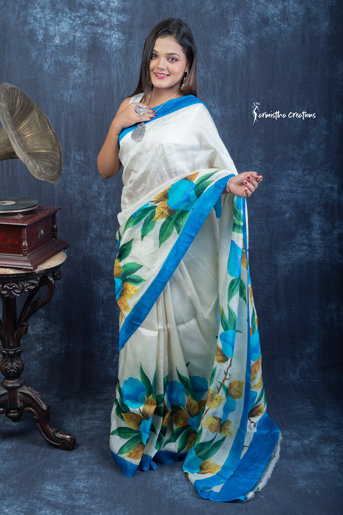 This floral hand painted Bishnupuri silk saree features vibrant blue blooms and mustard-gold leaves on a soft ivory base with a sky-blue border. Made from pure Bengal silk, it’s graceful, light, and perfect for radiant daywear.