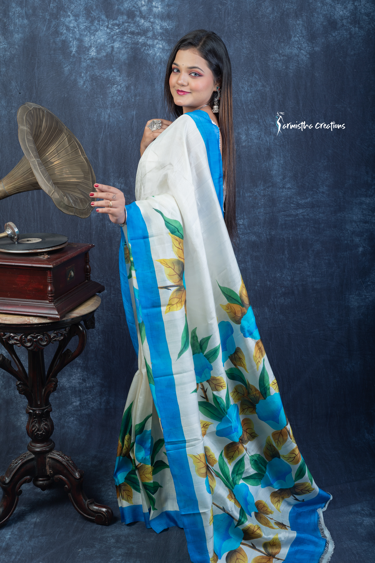 This floral hand painted Bishnupuri silk saree features vibrant blue blooms and mustard-gold leaves on a soft ivory base with a sky-blue border. Made from pure Bengal silk, it’s graceful, light, and perfect for radiant daywear.