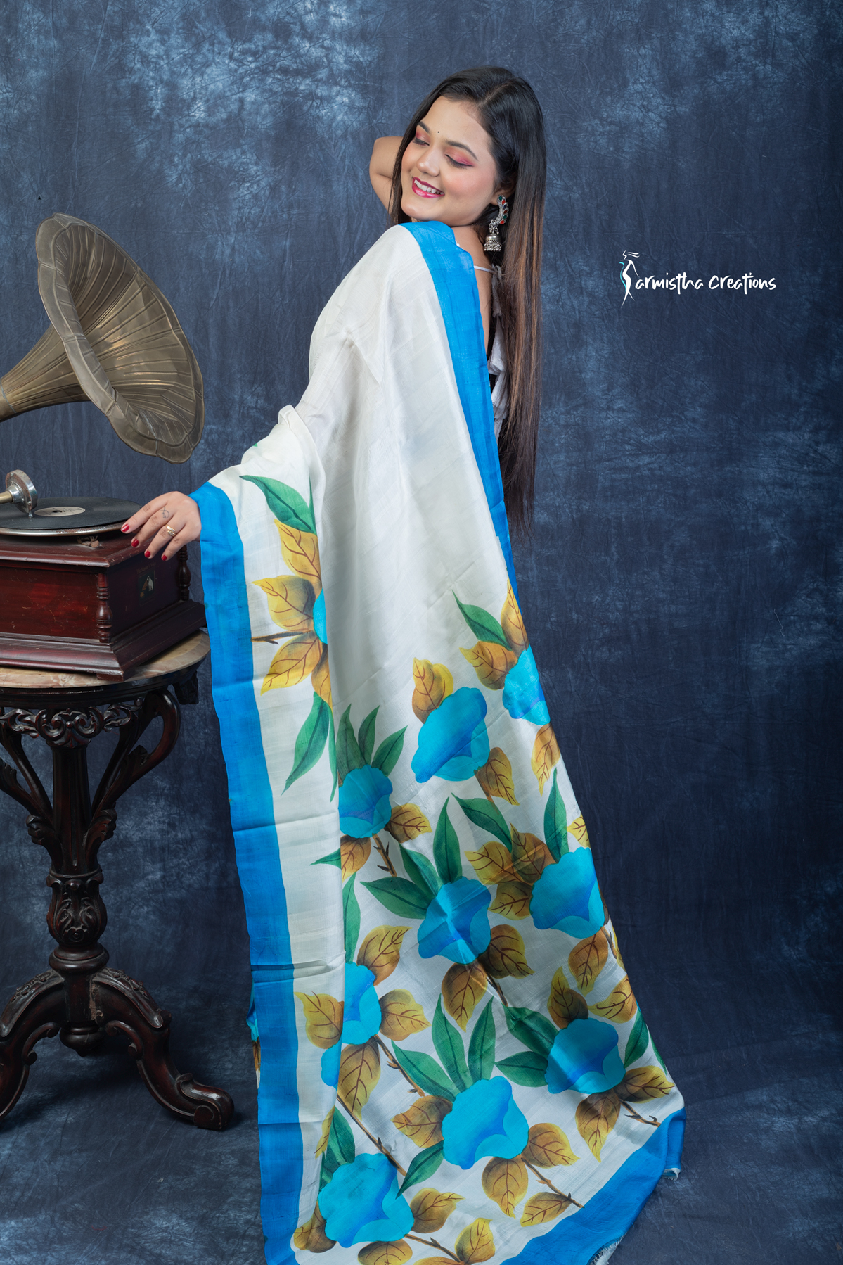 This floral hand painted Bishnupuri silk saree features vibrant blue blooms and mustard-gold leaves on a soft ivory base with a sky-blue border. Made from pure Bengal silk, it’s graceful, light, and perfect for radiant daywear.