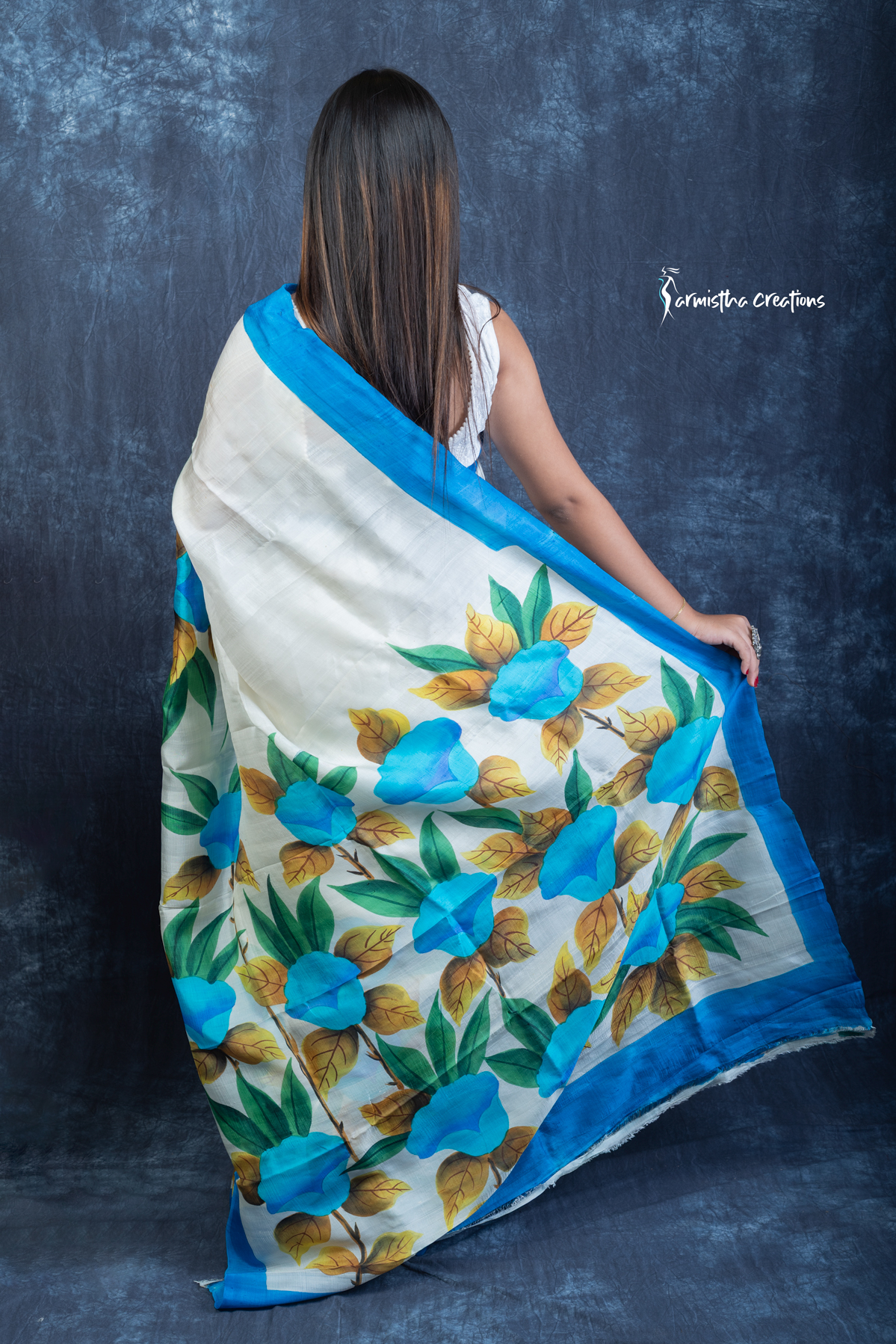 This floral hand painted Bishnupuri silk saree features vibrant blue blooms and mustard-gold leaves on a soft ivory base with a sky-blue border. Made from pure Bengal silk, it’s graceful, light, and perfect for radiant daywear.