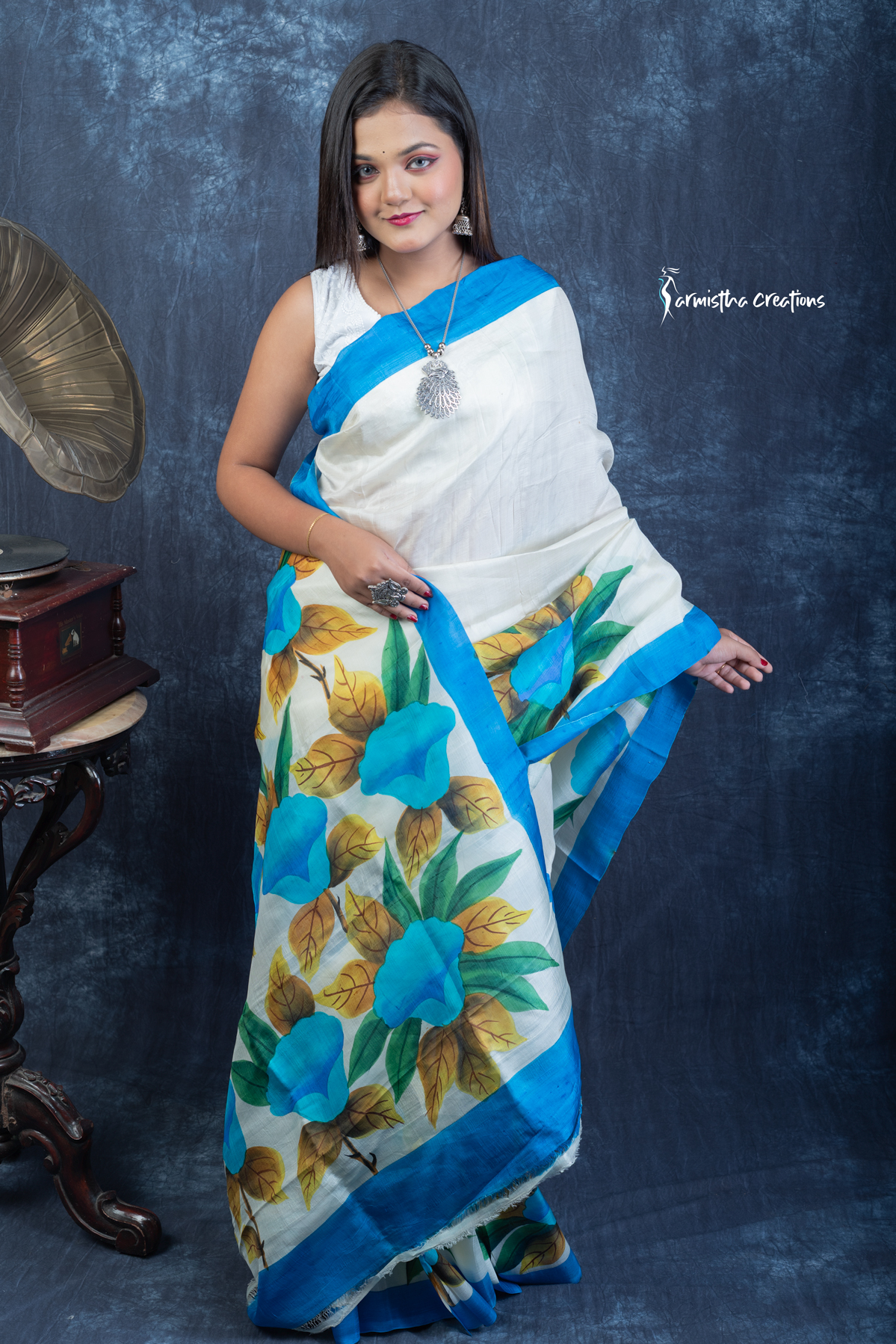 This floral hand painted Bishnupuri silk saree features vibrant blue blooms and mustard-gold leaves on a soft ivory base with a sky-blue border. Made from pure Bengal silk, it’s graceful, light, and perfect for radiant daywear.